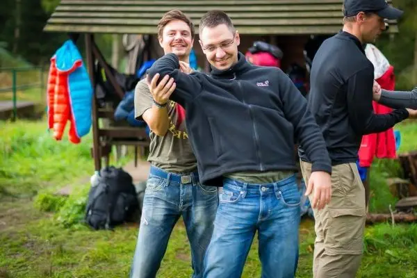 Drei Personen stehen in einer Wiese neben einer Holzunterkunft und interagieren miteinander