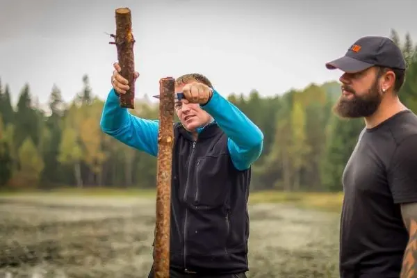 Mann bearbeitet Holzstück mit Messer, während ein anderer zuschaut