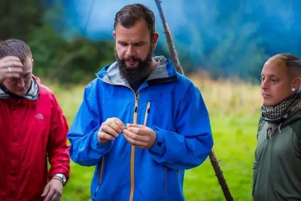 Mann in blauer Jacke hält einen kleinen Gegenstand in der Hand, zwei weitere Personen beobachten