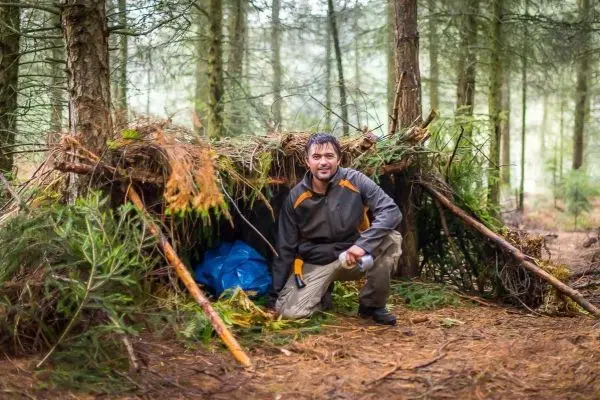 Mann kniet vor einer selbstgebauten Tarp-Notunterkunft im Wald