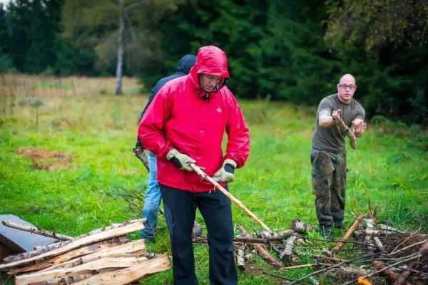 Mann mit rotem Regenmantel bearbeitet Holzstück mit Messer, im Hintergrund weitere Personen