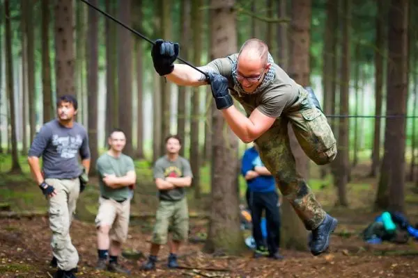 Mann überquert selbstgebaute Seilbrücke im Wald, Zuschauer beobachten