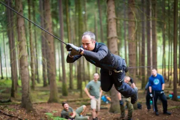 Mann überquert selbstgebaute Seilbrücke im Wald, Zuschauer im Hintergrund