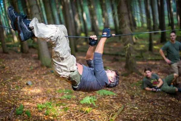 Mann überquert selbstgebaute Seilbrücke im Wald, zwei Personen beobachten
