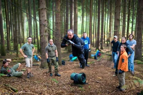 Mann überquert selbstgebaute Seilbrücke zwischen Bäumen, Gruppe beobachtet