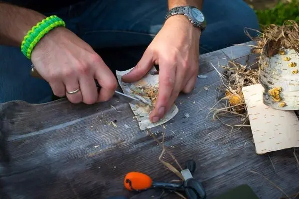 Messer schneidet Rinde auf einer Holzoberfläche, daneben liegen Zunder und Werkzeuge