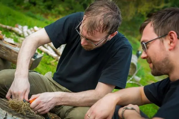 Messer schneidet Zunder auf einem Holzstück, während ein anderer zuschaut