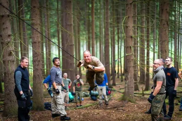 Person schwingt an Seil zwischen Bäumen, Gruppe beobachtet beim Survival Training