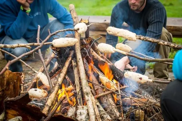 Personen sitzen um ein Feuer und rösten Teigstücke auf Stöcken beim Überlebenstraining