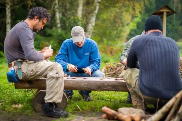 Vier Personen sitzen an einem Holzstamm und bearbeiten Materialien mit Messern