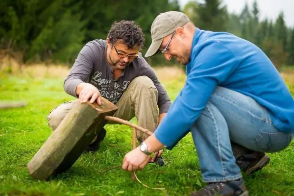 Zwei Personen bauen eine Schlagfalle mit einem Holzstück und einem Stock