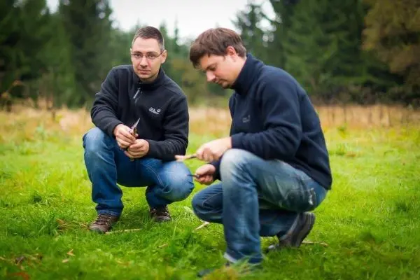 Zwei Personen knien auf einer Wiese und bearbeiten Holzstücke mit Messern