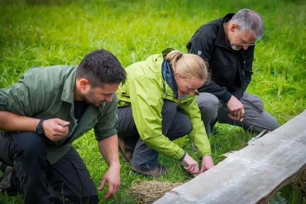 Drei Personen knien im Gras und bereiten Zunder auf einem Holzstück vor