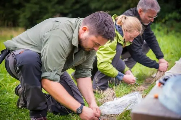 Drei Personen knien im Gras und binden Schnüre zusammen beim Überlebenstraining