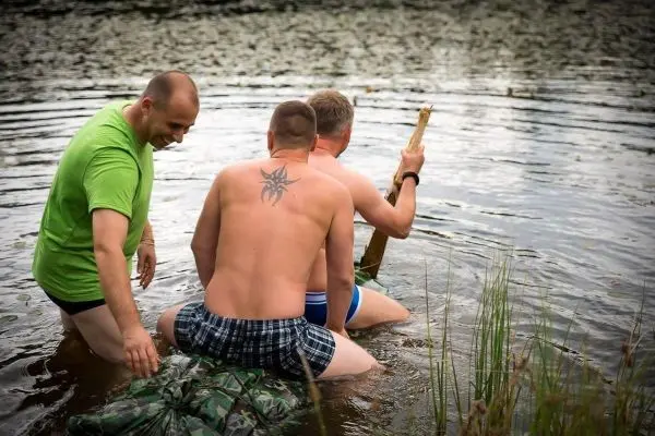 Drei Personen sitzen auf einer camouflagefarbenen Unterlage im Wasser und halten einen Stock