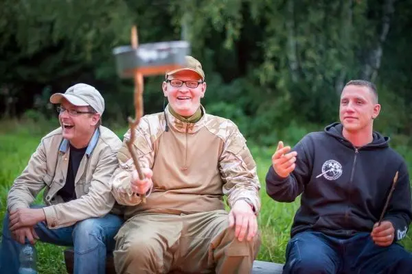 Drei Personen sitzen auf einer Holzbank und lachen, einer hält einen Stock mit einem Topf