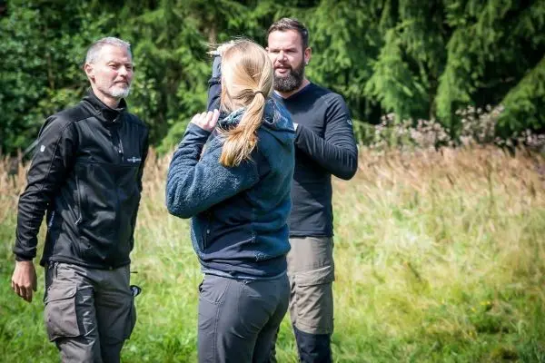 Drei Personen stehen in einer Wiese und interagieren miteinander