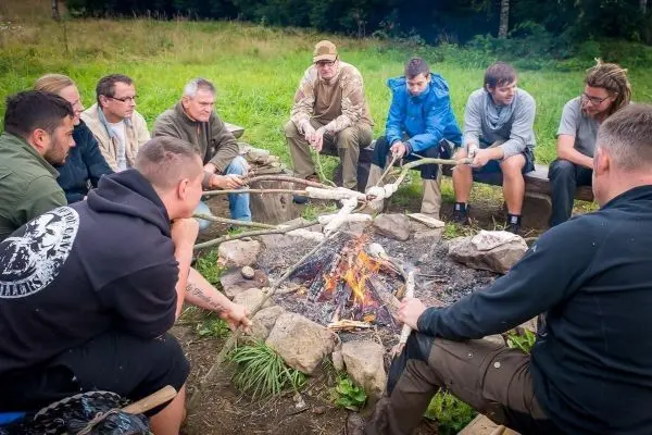 Gruppe sitzt um ein Feuer und hält Stöcke in der Hand beim Überlebenstraining