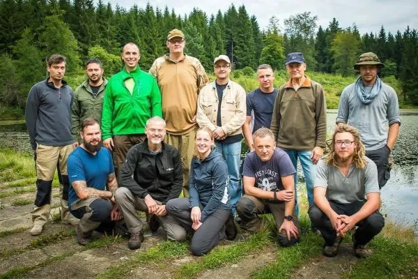 Gruppierung von Personen am Ufer eines Gewässers in bewaldeter Umgebung