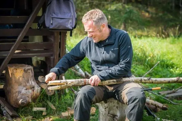 Holzstück wird mit einem Messer bearbeitet, während auf einem Baumstumpf gesessen wird
