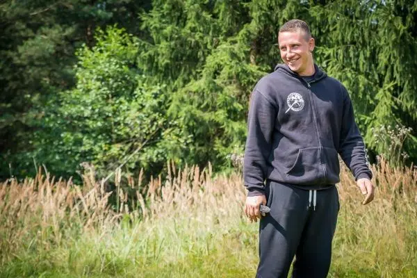 Lächelnde Person steht in einer Wiese mit hohem Gras und Bäumen im Hintergrund