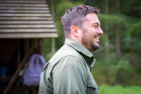 Lächelnde Person steht neben einer Holzstruktur im Wald