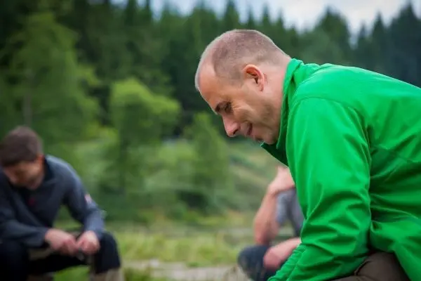 Mann in grüner Jacke beugt sich über den Boden, während andere Personen im Hintergrund sitzen