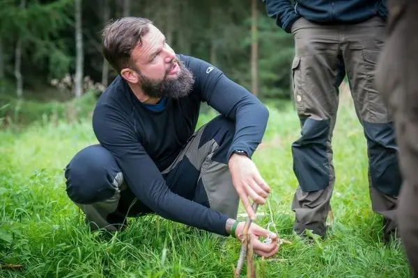 Mann kniet im Gras und bindet zwei Äste mit einer Schnur zusammen