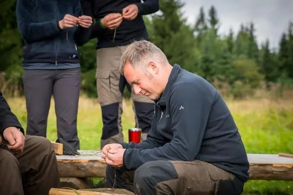 Mann knüpft mit beiden Händen an einem Tisch, im Hintergrund weitere Personen