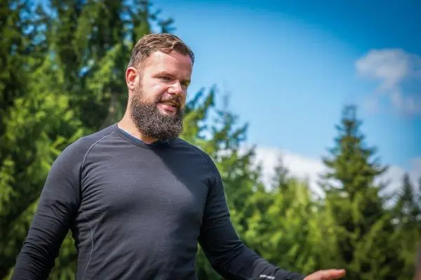 Mann mit Bart in schwarzem Shirt steht vor grünen Bäumen