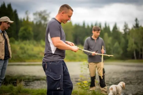 Mann schnitzt einen Stock am Ufer eines Gewässers, zwei weitere Personen im Hintergrund