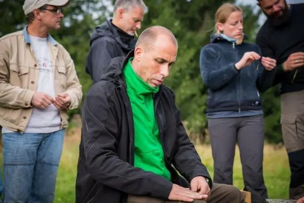 Mann sitzt auf einem Baumstamm und bearbeitet Holz mit einem Messer