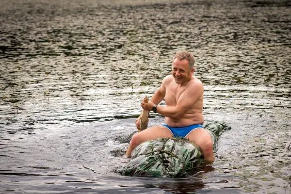 Mann sitzt auf einem camouflagefarbenen Objekt im Wasser und hält einen Stock