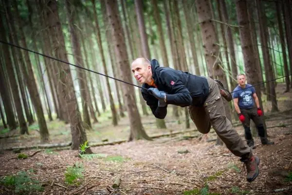 Mann überquert selbstgebaute Seilbrücke im Wald, während ein anderer zuschaut
