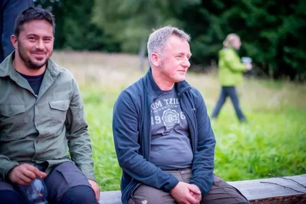 Zwei Männer sitzen auf einer Holzbank, im Hintergrund eine Person mit Kamera