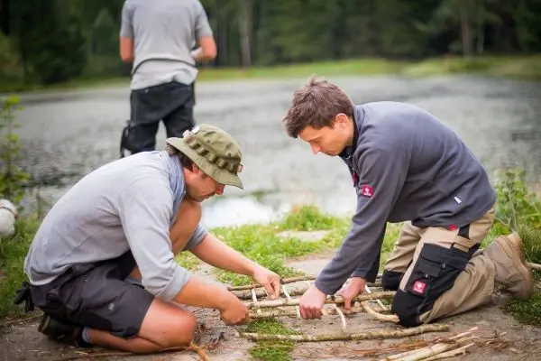 Zwei Personen bauen eine selbstgebaute Konstruktion aus Ästen am Ufer eines Gewässers
