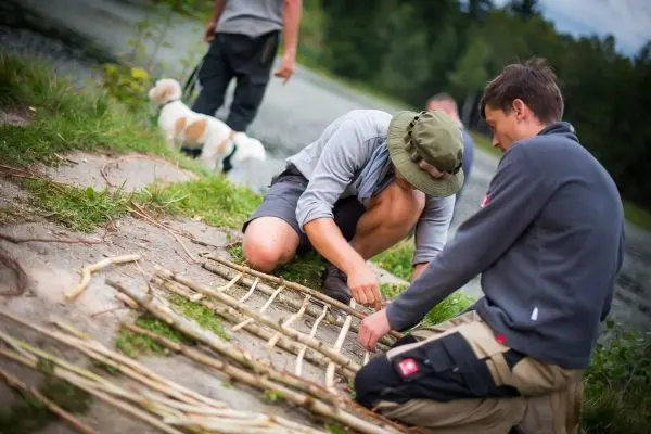 Zwei Personen bauen eine Struktur aus Ästen am Ufer eines Gewässers