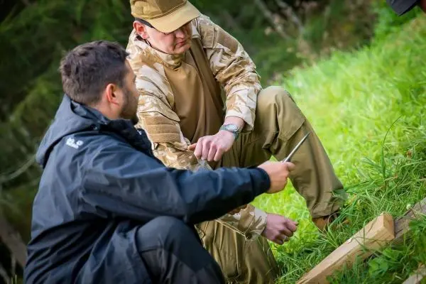 Zwei Personen knien im Gras und bearbeiten ein Holzstück mit einem Messer