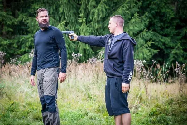 Zwei Personen stehen in einer Wiese, eine hält eine Pistole auf die andere