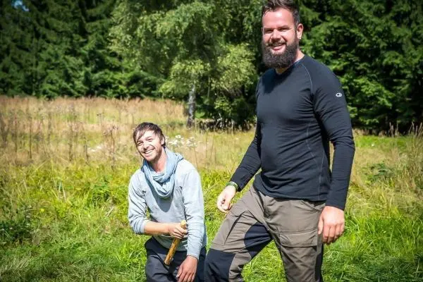 Zwei Personen stehen in einer Wiese, eine hält einen Stock in der Hand