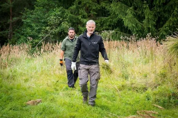 Zwei Personen tragen Gras und arbeiten in einer Wiese mit hohem Bewuchs