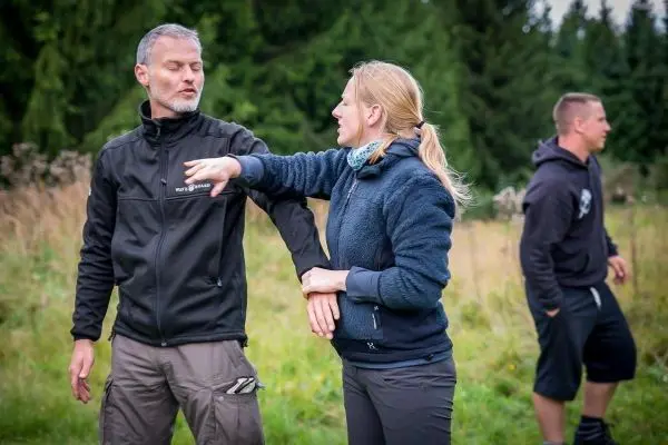 Zwei Personen üben eine Selbstverteidigungstechnik im Freien, während eine dritte Person im Hintergrund steht