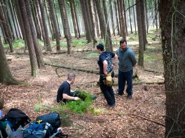 Drei Personen stehen und sitzen im Wald, eine Person untersucht den Boden