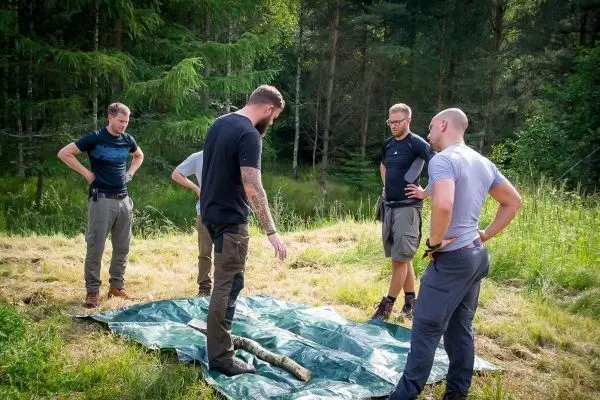 Fünf Personen stehen um eine Tarp-Notunterkunft und betrachten einen Stock