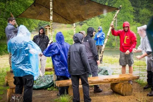Gruppe in Regenjacken steht unter einem Tarp, Holzbank im Vordergrund