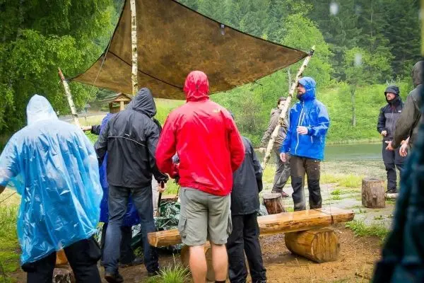 Gruppe in Regenjacken steht unter einer Tarp-Notunterkunft am Wasser