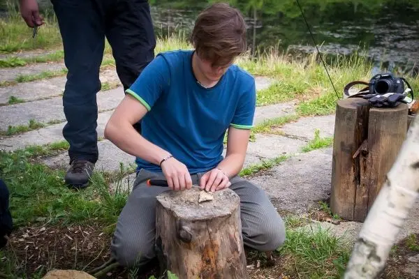 Junger Mann schnitzt mit einem Messer auf einem Holzstück am Ufer eines Gewässers