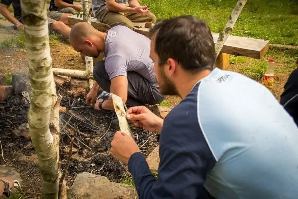 Mann bereitet Zunder am Bodenfeuer vor, während ein anderer Holzstücke bearbeitet