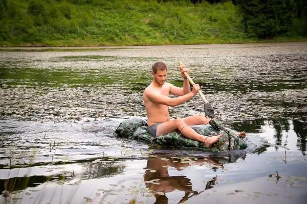 Mann paddelt mit einem Stock auf einer improvisierten Schwimmkonstruktion im Wasser