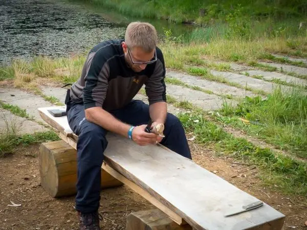 Mann schnitzt mit einem Messer an einem Holzstück auf einer Bank